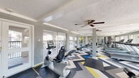 Located at 2400 Westheimer Rd in Houston's vibrant Montrose neighborhood, this fitness center offers a well-equipped exercise space designed for a comprehensive workout experience. The room is immediately distinguished by its bold, multi-toned chevron-patterned carpet in shades of gray, blue, yellow, and white, adding a dynamic visual energy to the space.

Natural light floods the interior through expansive floor-to-ceiling sliding glass windows and doors that line the walls, offering views of and direct access to an outdoor patio area featuring seating and lush plantings. Ceiling fans help maintain a comfortable environment throughout the room.

The fitness center is outfitted with a diverse range of equipment, including treadmills, stationary and recumbent bikes, an elliptical trainer, a multi-station cable weight machine, free weights, and adjustable benches. Full-length mirrors are mounted along one wall, enhancing the sense of space while allowing users to monitor their form during workouts.

The textured ceiling and neutral gray walls provide a clean, modern backdrop that keeps the focus on the fitness equipment. A support column near the center of the room is seamlessly integrated into the layout. The space also includes a fire safety station and posted exercise room rules, underscoring its well-maintained and safety-conscious design.