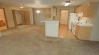 This single-floor space features an open-concept layout that seamlessly connects the living area and kitchen, creating a cohesive and functional environment. The kitchen is equipped with white appliances, including a refrigerator and electric range, complemented by light wood cabinetry. A peninsula with a built-in sink serves as the central feature of the kitchen, offering additional counter space and acting as a natural divider between the kitchen and the living area.

The living area is finished with neutral-toned carpet, providing a warm contrast to the hardwood-style flooring found in the kitchen and surrounding spaces. A chandelier hangs above the living area, adding a subtle decorative touch. Flush-mounted ceiling lighting in the kitchen ensures the workspace remains well-lit.

Natural wood-toned doors are visible throughout the space, contributing to the warm, cohesive aesthetic. A doorway visible toward the rear of the unit leads to what appears to be additional storage or a utility area. The open layout and neutral color palette throughout create a clean, inviting atmosphere well-suited for everyday living.