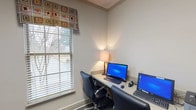 This business center is a compact, single-floor workspace designed to meet essential professional needs in a clean and functional environment. The room is finished with neutral gray walls and durable hard-surface flooring, creating a practical and focused atmosphere throughout.

The primary workstation area features a long, built-in desk surface accommodating two computer monitors, each paired with a full keyboard and mouse. Two black leather executive chairs provide comfortable seating at the shared workstation. A table lamp positioned on the desk surface adds warm ambient lighting to complement the natural light entering through a window dressed with horizontal blinds and a decorative geometric-patterned valance.

A Canon multifunction device serves as the center's printing and copying hub, positioned along one wall alongside additional built-in counter space and filing storage. A telephone and document organizers are also accessible within the space, supporting a range of administrative functions.

The business center is accessed through a distinctive glass-paned interior door featuring a multi-light grid design, allowing visibility into the space while maintaining a degree of separation. Overall, the business center offers a well-equipped, no-frills environment suited for productivity and professional tasks.