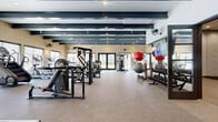 This modern fitness center offers a generously sized, single-floor layout designed to accommodate a comprehensive range of workout activities. The main exercise floor features durable, low-pile carpet and is lined with an extensive array of strength training machines, free weights, and cardio equipment, providing users with a full-body workout experience. Bold dark exposed beams run the length of the ceiling, lending an industrial-chic aesthetic that contrasts cleanly with the light walls and warm brick accents. Large windows and glass double doors along the perimeter flood the space with natural light while offering views of the surrounding outdoor landscape. Wall-mounted fans and flat-screen televisions are thoughtfully positioned throughout for comfort and entertainment during workouts. A dedicated studio space adjacent to the main floor features rich hardwood-style flooring, textured stone-tile walls, full-length mirrors, interactive fitness displays, and additional wall-mounted fans — ideal for yoga, stretching, or group fitness classes. A large flat-screen television is mounted within a recessed niche in the studio, with built-in storage below for exercise balls and accessories. The overall ambiance is clean, modern, and energizing, combining functional design with refined architectural details.