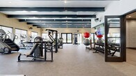 This modern fitness center offers a generously sized, single-floor layout designed to accommodate a comprehensive range of workout activities. The main exercise floor features durable, low-pile carpet and is lined with an extensive array of strength training machines, free weights, and cardio equipment, providing users with a full-body workout experience. Bold dark exposed beams run the length of the ceiling, lending an industrial-chic aesthetic that contrasts cleanly with the light walls and warm brick accents. Large windows and glass double doors along the perimeter flood the space with natural light while offering views of the surrounding outdoor landscape. Wall-mounted fans and flat-screen televisions are thoughtfully positioned throughout for comfort and entertainment during workouts. A dedicated studio space adjacent to the main floor features rich hardwood-style flooring, textured stone-tile walls, full-length mirrors, interactive fitness displays, and additional wall-mounted fans — ideal for yoga, stretching, or group fitness classes. A large flat-screen television is mounted within a recessed niche in the studio, with built-in storage below for exercise balls and accessories. The overall ambiance is clean, modern, and energizing, combining functional design with refined architectural details.