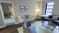 This thoughtfully designed single-floor space exudes a refined, contemporary aesthetic that balances comfort with sophistication. The living and dining areas flow seamlessly together, creating an open and welcoming atmosphere ideal for both relaxing and entertaining.

The dining area features a round glass-top table with sleek chrome legs, paired with clean-lined white chairs that contribute to the space's modern sensibility. Above, a pair of black-framed black-and-white photographs add a touch of urban artistry to the soft gray walls. Rich hardwood flooring runs throughout the space, providing warmth and grounding the overall design.

The living area is anchored by a plush, curved gray accent chair set atop a light green area rug, offering a cozy and stylish seating vantage point. A glass coffee table and a compact side desk with a floor lamp complete the arrangement, providing both functional workspace and ambient lighting. Natural light filters in through a window, brightening the room and enhancing its airy feel.

Elegant crown molding along the ceiling adds an architectural detail that elevates the space, lending it a classic character that complements its modern furnishings. The overall ambiance is calm, polished, and inviting — a well-appointed space designed for comfortable everyday living.