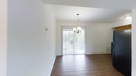 This single-floor residence offers a clean, well-maintained interior defined by a bright and open aesthetic. The main living and dining area is grounded by rich, dark hardwood flooring that adds warmth and contrast to the crisp white walls and ceiling. A classic chandelier with candelabra-style arms serves as a focal point overhead, adding a touch of elegance to the space. Natural light flows generously through a large sliding glass door fitted with vertical blinds, which opens to an outdoor area — likely a balcony or patio — where lush greenery is visible beyond the railing. This seamless connection between the interior and exterior creates an airy, inviting ambiance throughout. The walls are finished in a neutral white palette, lending a versatile and timeless backdrop suitable for a variety of furnishing styles. The space is thoughtfully laid out, with a smooth transition between living areas. Overall, the residence presents a bright, practical, and welcoming environment, balancing functional design with tasteful details that make it comfortable and easy to personalize.
