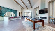 This single-story amenity lounge is designed to impress, combining architectural drama with thoughtfully curated social spaces. Soaring ceilings with exposed wood beams define the open main area, where abundant natural light pours in through floor-to-ceiling windows and transom glazing. A central pool table anchors the room atop a layered area rug, while a shuffleboard table positioned near expansive windows offers views of the surrounding neighborhood and outdoor pool area.

The entertainment zone features a linear fireplace clad in white tile, flanked by vertical wood slatted wall panels and a wall-mounted television. Curated abstract artwork and warm accent lighting contribute to the refined yet relaxed atmosphere. A dedicated seating area with lounge chairs and glass-topped dining tables sits adjacent to double doors that open directly to the outdoor pool terrace.

The bar and kitchen area is a standout feature, showcasing sage green cabinetry paired with striking teal glazed tile backsplash and a dramatic arched mirror detail. Marble-look countertops, dual sinks, stainless steel appliances, and built-in open shelving complete this fully equipped social kitchen. Wood-look plank flooring flows throughout, unifying the space with a warm, contemporary finish.
