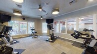 Located at 16432 Fox Hunt Dr in Strongsville, Ohio, this single-level fitness center offers a thoughtfully designed workout environment suited for a range of cardio and light strength training activities. The space is outfitted with multiple Matrix-brand cardio machines, including recumbent bikes, upright cycles, spin bikes, and ellipticals, all arranged to allow comfortable circulation throughout the room.

The interior features neutral-toned walls accented by crisp crown molding, and the floor is covered in a patterned modular carpet tile that provides both durability and a polished aesthetic. Full-length wall mirrors run along one side of the room, effectively expanding the visual scale of the space while allowing users to monitor their form during workouts. Multiple wall-mounted flat-screen televisions are positioned for easy viewing from various equipment stations.

Natural light filters in through large windows on multiple walls, offering views of an outdoor deck and surrounding greenery. Ceiling fans with integrated light fixtures provide supplemental air circulation and lighting alongside flush-mounted ceiling fixtures. The textured ceiling adds a traditional finish consistent with the building's overall style. Adjacent interior windows connect the fitness room to a neighboring common area, contributing to an open, community-oriented atmosphere.