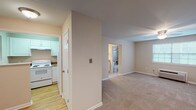 This single-floor studio is efficiently designed to maximize functionality within a compact footprint. The open main living area is finished with wall-to-wall carpet and features a ceiling fan with integrated lighting, a wall-mounted HVAC unit, and a window that brings in natural light, creating a comfortable and airy atmosphere. Neutral-toned walls throughout contribute to a clean, versatile aesthetic.

The separate kitchen is distinctly defined from the living space and features hardwood-style flooring, white cabinetry, stone-patterned countertops, a double-basin stainless steel sink, and a white electric coil range with an overhead exhaust hood. A window with blinds provides additional ventilation and light to the cooking area.

The full bathroom includes a white vanity with stone-patterned countertop, a framed mirror, and a tiled tub-shower combination with a window above for natural light. Tile flooring and brushed nickel fixtures complete the space with a polished, cohesive finish.

A central dividing wall with a paneled door separates the living and utility areas, adding a degree of privacy and organization. Overall, the studio offers a practical, move-in-ready layout suited for efficient single-occupant living.