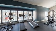 This fitness center is thoughtfully designed to accommodate a full range of workout activities across a single, open floor. The space is divided into distinct functional zones, each tailored to specific exercise needs. The strength training area features a multi-station cable machine, a weight bench with free weights, and a vertical knee raise tower, all set against deep teal accent walls and floor-to-ceiling mirrors that amplify the sense of space. Large windows with slatted blinds allow ample natural light to filter in while maintaining privacy. Adjacent to the strength zone, a dedicated cardio section houses multiple treadmills and additional cardio equipment positioned facing outward-facing windows, providing views to the exterior. The expansive group exercise studio offers an open, unobstructed layout with high vaulted ceilings, ceiling fans for air circulation, wall-mounted mirrors, a ballet-style barre counter, and a full rack of fitness accessories including resistance bands, medicine balls, stability balls, and foam rollers. Recessed and mounted lighting complement the natural light throughout. A wall-mounted flat-screen television is positioned for visibility across the main floor. Durable rubber flooring runs throughout the entire facility, providing a safe and practical surface for all exercise activities. The overall ambiance is clean, modern, and functionally organized.