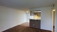 This single-floor residence offers a straightforward and functional layout centered around an open living area. The living space is finished with plush dark carpeting that extends throughout, providing a warm and comfortable underfoot feel. A ceiling fan is centrally positioned overhead, ensuring year-round air circulation, while a textured popcorn ceiling adds a classic residential character to the room. Vertical blinds along the window offer privacy and light control, allowing natural light to filter softly into the space.

A defining architectural feature is the pass-through kitchen, which connects to the living area via a wood-paneled breakfast bar complete with an open lattice partition above. This design element creates a visual divide while maintaining an open and connected feel between the two areas. The kitchen is equipped with light wood cabinetry and a white stovetop range, offering a clean and utilitarian cooking environment. The countertop along the bar provides casual seating and dining opportunities, making the space well-suited for everyday living. The overall ambiance is practical and uncluttered, with a neutral color palette of whites and warm browns throughout.
