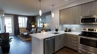 This single-floor space showcases a thoughtfully designed open-concept layout that seamlessly connects the kitchen and living area. The kitchen is a standout feature, equipped with warm wood-toned cabinetry that pairs elegantly with crisp white quartz countertops. A peninsula with an undermount sink serves as both a functional workspace and a natural divider between the kitchen and living space. Stainless steel appliances, including a dishwasher, add a polished, contemporary touch. A white subway tile backsplash completes the clean aesthetic. Two sleek elongated pendant lights hang above the peninsula, providing focused task lighting while enhancing the modern design. Upper and lower cabinetry along the perimeter offers generous storage. The open living area beyond features dark hardwood flooring, creating warmth and contrast against the lighter kitchen elements. A large window allows natural light to filter into the space, illuminating a cozy seating arrangement with layered textiles and greenery. The overall ambiance is modern yet inviting, balancing functionality with contemporary style throughout.
