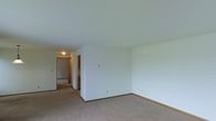 This single-floor space presents a clean, functional layout highlighted by neutral tones throughout. The main living area features wall-to-wall carpeting in a warm beige tone, providing a comfortable and cohesive foundation. Smooth white walls and a standard flat ceiling create a bright, airy atmosphere, while the understated color scheme offers versatility for a variety of furnishing styles.

A central hallway connects the main living area to additional rooms beyond, with warm wood-toned door frames and trim providing subtle contrast against the white walls. The hallway is illuminated by a ceiling-mounted light fixture, ensuring consistent visibility throughout the passage. Glimpses of adjacent spaces, including what appears to be a kitchen area distinguished by cabinetry with warm wood finishes, are visible through the corridor.

Electrical outlets are conveniently positioned along the walls, and light switch panels are accessible near key entry points. The consistent use of wood-toned baseboards throughout ties the spaces together cohesively. Overall, the unit offers a practical and move-in-ready environment with a straightforward, functional design.