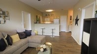 This single-floor residence showcases an open-concept layout that combines the living and kitchen areas into one cohesive, flowing space. The living area is anchored by a generously sized sectional sofa accented with decorative throw pillows, paired with a glass-top coffee table that adds a touch of elegance. Warm hardwood-style flooring runs throughout, complementing the soft, neutral wall tones that create a welcoming ambiance.

The kitchen is positioned behind a raised breakfast bar, which accommodates two sleek modern bar stools, making it an ideal spot for casual dining or entertaining. Light wood cabinetry lines the kitchen walls, offering ample storage, while white appliances including a refrigerator and microwave provide a clean, classic aesthetic. The under-cabinet lighting adds a practical yet warm glow to the workspace.

A ceiling-mounted light fixture illuminates the open area, contributing to the bright and airy feel of the space. Multiple doorways visible from the main living area suggest additional rooms, including what appears to be a hallway leading to further living quarters. The overall design balances functionality with comfort, making it a practical and inviting living environment.