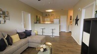 This single-floor residence showcases an open-concept layout that combines the living and kitchen areas into one cohesive, flowing space. The living area is anchored by a generously sized sectional sofa accented with decorative throw pillows, paired with a glass-top coffee table that adds a touch of elegance. Warm hardwood-style flooring runs throughout, complementing the soft, neutral wall tones that create a welcoming ambiance.

The kitchen is positioned behind a raised breakfast bar, which accommodates two sleek modern bar stools, making it an ideal spot for casual dining or entertaining. Light wood cabinetry lines the kitchen walls, offering ample storage, while white appliances including a refrigerator and microwave provide a clean, classic aesthetic. The under-cabinet lighting adds a practical yet warm glow to the workspace.

A ceiling-mounted light fixture illuminates the open area, contributing to the bright and airy feel of the space. Multiple doorways visible from the main living area suggest additional rooms, including what appears to be a hallway leading to further living quarters. The overall design balances functionality with comfort, making it a practical and inviting living environment.