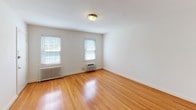 Located at 7025 Strathmore St, Building 333, Unit 3 in Chevy Chase, Maryland, this single-floor residence presents a clean, functional layout with a blend of classic and updated finishes throughout. The living areas feature warm-toned hardwood flooring that extends through multiple rooms, lending a cohesive and inviting feel to the space. Large windows in several rooms allow natural light to filter in, offering views of surrounding greenery and creating a bright, airy atmosphere. A wall-unit HVAC system and classic cast-iron radiators provide year-round climate control. The kitchen has been thoughtfully updated with honey-toned wood cabinetry, granite countertops, a stainless steel sink, and a stainless steel refrigerator, all set atop tile flooring. The bathroom retains a timeless character with white subway tile surrounding a full soaking tub and shower combination, classic hex-tile flooring, and a bright window that adds natural ventilation. One carpeted room, bathed in natural light from a bank of windows overlooking lush trees, offers a comfortable, quiet retreat. Neutral white walls throughout the unit provide a versatile backdrop, making the space easy to personalize. The overall ambiance is calm and well-maintained, well-suited to those seeking a straightforward, comfortable residence in a desirable Maryland neighborhood.