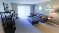 This single-floor space offers a comfortable and stylishly designed living environment. The living room serves as the centerpiece, showcasing a spacious layout with plush wall-to-wall carpeting that adds warmth underfoot. A large sliding glass door fitted with vertical blinds floods the room with natural light while providing access to the outdoors. The neutral gray walls create a calm, modern backdrop, complemented by a set of decorative circular mirrors that add a contemporary touch to the space. The room is furnished with a substantial dark leather sectional sofa and a matching tufted ottoman, offering ample seating and a cohesive aesthetic. Accent pillows in bold red and patterned tones introduce a pop of color, enhancing the room's visual interest. A media console along the adjacent wall provides practical storage and entertainment options. The overall ambiance is relaxed yet polished, blending functional furniture choices with thoughtful decorative details. The generous proportions of the living area allow for easy movement and flexible furniture arrangements, making this a welcoming and versatile space well-suited for both everyday living and casual entertaining.