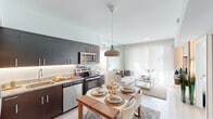 Located at 15370 SW 136th St in Miami, FL, this single-floor residence offers a thoughtfully designed open-concept layout filled with natural light. The kitchen is a standout feature, equipped with dark espresso cabinetry, granite countertops, a classic subway tile backsplash with under-cabinet lighting, and a full suite of stainless steel appliances. A pendant light anchors the adjacent dining area, which flows seamlessly into the bright living room. The living space is highlighted by large floor-to-ceiling windows dressed in sheer white curtains, a textured wood-panel accent wall, and light hardwood-style flooring that runs throughout the main areas. The primary bedroom is a serene retreat with soft neutral tones, a wide window for natural light, and a clean, minimalist aesthetic. A second bedroom functions effectively as a flexible space, featuring built-in shelving and a dedicated desk area. Both bathrooms are finished with large-format tile flooring, granite vanities with dark wood cabinetry, and tub-shower combinations. The overall ambiance of the residence is modern and airy, combining functional design with warm, contemporary finishes ideal for comfortable urban living in Miami.