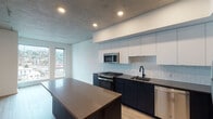 Located in the heart of Portland's west side at 1510 SW Alder St, this single-floor unit showcases a sleek, modern design with thoughtful finishes throughout. The open-concept kitchen is a standout feature, highlighted by dark navy lower cabinetry contrasted with crisp white upper cabinets, a soft tile backsplash, and dark quartz countertops extending across a spacious island. Stainless steel appliances, including a gas range, microwave, dishwasher, and refrigerator, complete the well-appointed cooking space. Light hardwood flooring flows seamlessly through the main living areas, adding warmth against the exposed concrete ceiling that lends an industrial character to the space. Natural light filters in through windows that offer views of the surrounding Portland cityscape. The bathroom is elegantly finished with large-format marble-look tile surrounding the soaking tub and shower, complemented by dark cabinetry, a gray quartz vanity top, and matte black fixtures throughout. A generously sized walk-in closet with wire shelving and carpeted flooring provides ample storage. The overall ambiance balances urban industrial elements with refined modern finishes, creating a polished and functional living environment.
