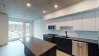 Located in the heart of Portland's west side at 1510 SW Alder St, this single-floor unit showcases a sleek, modern design with thoughtful finishes throughout. The open-concept kitchen is a standout feature, highlighted by dark navy lower cabinetry contrasted with crisp white upper cabinets, a soft tile backsplash, and dark quartz countertops extending across a spacious island. Stainless steel appliances, including a gas range, microwave, dishwasher, and refrigerator, complete the well-appointed cooking space. Light hardwood flooring flows seamlessly through the main living areas, adding warmth against the exposed concrete ceiling that lends an industrial character to the space. Natural light filters in through windows that offer views of the surrounding Portland cityscape. The bathroom is elegantly finished with large-format marble-look tile surrounding the soaking tub and shower, complemented by dark cabinetry, a gray quartz vanity top, and matte black fixtures throughout. A generously sized walk-in closet with wire shelving and carpeted flooring provides ample storage. The overall ambiance balances urban industrial elements with refined modern finishes, creating a polished and functional living environment.