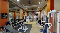 This fitness center is thoughtfully designed to support a full range of workout routines within a single, open-plan space. The room features a tray ceiling with recessed lighting and multiple ceiling fans, ensuring consistent airflow and a well-lit environment throughout. Warm tan walls are accented by bold orange columns, creating an energizing aesthetic that defines the space visually.

Large floor-to-ceiling mirrors are positioned along the walls, framing the orange accent columns and visually expanding the room while allowing users to monitor their form during exercise. Multiple windows along the perimeter allow natural light to filter in, providing views of the surrounding outdoor greenery.

The fitness center is comprehensively outfitted with a wide array of white-framed strength machines, including cable systems, leg press, and various isolation machines. A full dumbbell rack with an extensive range of weights occupies a central position, accompanied by adjustable benches and rubber floor mats to define the free weights area. Additional functional fitness equipment includes medicine balls, a stability ball, a ball rack, and exercise mats. A wall-mounted television is installed for entertainment during workouts. The combination of organized equipment layout, ample natural light, and a vibrant design scheme creates a motivating and functional fitness environment.