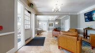 This well-appointed lobby and common area presents a polished, professional aesthetic rooted in classic architectural design. The space is unified by large-format travertine-style tile flooring that extends throughout, lending warmth and a sense of openness to every area. Crown molding, white chair-rail wainscoting, and coffered ceiling details add refined character to the soft gray walls, creating an inviting yet sophisticated atmosphere.

The main seating area is furnished with a set of rich cognac leather armchairs arranged atop a patterned area rug, offering a comfortable gathering space for visitors. An elegant multi-arm chandelier anchors the ceiling overhead, complemented by modern wall sconces that provide warm, ambient lighting. A wall-mounted flat-screen display is positioned prominently for easy viewing.

Toward the rear of the space, a staffed reception window with plantation shutters serves as the main point of contact, flanked by a private office door. A secondary seating nook with a gray upholstered chair provides a quieter area for waiting. The entry vestibule features classic paneled glass doors and a welcome mat, transitioning seamlessly into the main lobby. A hallway leads further into the building, connecting this common area to additional interior spaces.