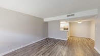 This single-floor unit offers a clean, functional layout designed for comfortable everyday living. The living area is open and airy, finished with wood-style laminate flooring in a neutral gray tone that extends throughout the entire unit. Textured ceilings and soft gray walls create a consistent, understated palette, while a large window near the front entry allows natural light to fill the space. Overhead HVAC vents are integrated into the ceiling, ensuring efficient climate control.

The kitchen is designed in a galley configuration, featuring white cabinetry with brushed hardware, light countertops, a double basin sink, a dishwasher, an electric range, and a full-size refrigerator. A pass-through opening connects the kitchen to the adjoining dining or living area, which is accented by an exposed brick wall, adding a distinctive architectural detail to the space.

The bedroom is well-sized with a window providing natural light and direct access to a walk-in closet equipped with built-in shelving. The bathroom includes a single-sink vanity with a speckled countertop, a large mirror, white cabinetry, and a tiled shower-tub combination, all maintained in a bright, clean finish.