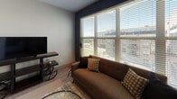 This single-floor unit showcases a contemporary urban design with industrial accents visible throughout, including exposed ductwork and metal hardware that lend an edgy, loft-inspired character. The kitchen is well-appointed with dark espresso cabinetry, stainless steel appliances including a French door refrigerator, built-in microwave, dishwasher, and a freestanding range topped by a stainless steel hood. Light quartz countertops and glass-front upper cabinets provide visual contrast, while wood-look flooring ties the space together seamlessly from the entry door through the kitchen and into the living area.

The living room benefits from oversized windows that invite natural light and offer views of the surrounding urban landscape. The bedroom is separated from the living space by a distinctive industrial-style sliding door system, adding both functionality and a design focal point. Soft carpeting and warm ambient lighting create a relaxed atmosphere in the bedroom. The bathroom features tile flooring, a dark vanity with a vessel sink, a framed mirror, and a glass-enclosed walk-in shower. A dedicated laundry area with a stacked washer and dryer and a fold-down utility shelf completes this thoughtfully designed unit.