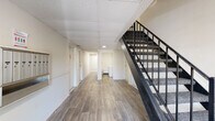 This shared common area serves as the central hub of a multi-unit residential building, designed to provide functional and accessible amenities for its residents. The space is finished with durable wood-look laminate flooring throughout, lending a clean and cohesive aesthetic to the corridors and common zones.

The main entrance lobby is well-defined, featuring a glazed entry door that allows natural light to filter into the space, creating a welcoming first impression. Adjacent to the entrance, a row of individual metal mailboxes is mounted along the wall, accompanied by a community bulletin board for resident notices and information. The lobby also includes a dark-toned entry mat for practicality.

A prominent staircase with dark metal railings and carpeted treads connects the floor to upper levels, serving as a central architectural element. The suspended tile ceiling is consistent throughout, fitted with recessed and flush-mounted lighting to ensure the corridors remain well-lit.

Toward the rear of the common area, a dedicated laundry room houses washer and dryer units, offering convenient on-site laundry access for residents. Multiple unit entry doors line the hallway, each clearly numbered. The overall environment is clean, neutral, and efficiently organized.