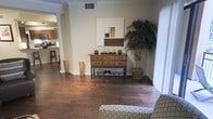 This contemporary living area showcases an open floor plan, seamlessly connecting different functional spaces. The room features rich, dark hardwood flooring that adds warmth and elegance to the space. A rustic wooden console table serves as a focal point, adorned with decorative items and flanked by tall, cylindrical vases, blending modern and traditional elements.

The space is brightened by natural light filtering through vertical blinds, hinting at a patio or balcony beyond. A large artificial plant in the corner brings a touch of nature indoors, softening the overall aesthetic. The walls are painted in a neutral tone, creating a calm backdrop for the furnishings.

A partial view of the kitchen area is visible, suggesting an open-concept design that promotes flow and connectivity between living spaces. The room's decor is minimalist yet thoughtful, with a geometric wall art piece adding visual interest. A comfortable armchair with a circular pattern provides a cozy seating option.

The ceiling features crown molding, adding a touch of architectural sophistication. Overall, the space exudes a harmonious blend of contemporary style and homey comfort, ideal for both relaxation and entertaining.