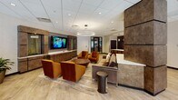Located at 2500 Clarendon Blvd in Arlington, Virginia, this single-floor amenity lounge is a well-appointed communal space designed for both relaxation and collaboration. The interior is finished with light wood-look flooring, neutral walls, and a drop ceiling with recessed lighting throughout, creating a bright and welcoming atmosphere.

The main lounge area features multiple seating configurations, including rich amber-toned velvet armchairs, a plush sofa, and a circular coffee table, all centered around a wall-mounted flat-screen TV set against textured, dark wood-paneled cabinetry. A round dining table with upholstered chairs provides an additional gathering spot, complemented by bold abstract artwork adorning the walls.

A fully equipped kitchenette anchors one end of the space, outfitted with dark cabinetry, a full-size refrigerator, a sink with a white countertop island, pendant lighting, and its own mounted TV — ideal for catered events or casual use.

At the opposite end, a large communal table seats many guests beneath a pair of striking circular pendant lights. Floor-to-ceiling glass windows and doors frame views of an outdoor patio and courtyard, flooding the space with natural light. Black-framed glass entry doors provide a polished transition from adjoining areas, completing the space's refined, contemporary aesthetic.
