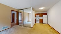 This single-floor residential unit offers a functional and straightforward layout designed for comfortable everyday living. The open living area flows directly into the kitchen, creating a cohesive shared space. The kitchen is equipped with warm-toned medium oak cabinetry, white appliances including an electric range and a full-sized refrigerator, a range hood, and light-toned countertops, all set on easy-care vinyl flooring. A notable feature of the living area is a set of full-length mirrored sliding closet doors, which visually expand the space and add a reflective brightness to the room. Soft carpet underfoot extends through the living and bedroom areas, contributing to a cozy, residential feel. The bedroom is generously sized, bathed in natural light through a wide sliding window that frames a view of surrounding greenery, and features textured ceiling and clean white walls with wood-trimmed accents. The bathroom is neatly appointed with white square wall tile surrounding a full soaking tub and shower, mosaic tile flooring, a wood-framed vanity with a wide countertop sink, and a large mirror. Consistent wood trim throughout the unit ties the spaces together with a warm, classic touch.