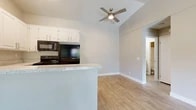 Located at 1440 Barton Rd in Redlands, California, this one-bedroom, one-bathroom residence offers a thoughtfully arranged single-floor layout. The combined kitchen and living area forms the heart of the unit, characterized by vaulted ceilings that lend a sense of openness beyond the unit's compact footprint. Light wood-style flooring runs throughout the main living spaces, complementing the neutral wall tones and clean white baseboards.

The kitchen is well-equipped with white cabinetry extending to the elevated ceiling line, granite-style speckled countertops, a peninsula breakfast bar, a black refrigerator, black range, dishwasher, and an integrated sink. A ceiling fan with lighting anchors the living area, which opens directly onto a balcony through sliding glass doors, providing outdoor views of a pool and surrounding greenery.

A hallway connects the main living area to the bedroom and bathroom. The bedroom features plush carpeting, a ceiling fan, a window with natural light, and access to a closet. The bathroom includes a white vanity with matching granite-style countertop, a toilet, and a tub-shower combination with tile surround and wood-look flooring, completing the unit's cohesive, contemporary aesthetic.