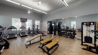 Located at 201 River Pointe Dr in Conroe, Texas, this single-level fitness center is well-equipped to support a wide range of workout routines. The space features durable wood-look flooring throughout, complemented by light gray walls and high ceilings that contribute to an open, airy atmosphere. Natural light flows in through multiple white-trimmed windows fitted with blinds, keeping the environment bright and inviting.

The cardio section is outfitted with treadmills, elliptical trainers, a stationary bike, and a rowing machine, along with a wall-mounted flat-screen television for entertainment during workouts. Ceiling fans and overhead fluorescent lighting ensure comfort and visibility throughout the space.

The strength training area is thoroughly equipped, featuring a cable machine, multiple selectorized strength machines, adjustable weight benches, and a full rack of dumbbells. An additional section is dedicated to functional fitness, housing medicine balls, resistance bands, stability balls, and foam rollers organized neatly on racks. Full-length mirrors line one wall, allowing users to monitor their form during exercises.

French doors and standard entry doors provide access to the facility, while informational postings and fitness charts are displayed on the walls. Overall, the fitness center offers a clean, organized, and fully equipped environment suited for a comprehensive workout experience.