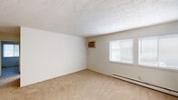 This single-floor unit features a straightforward, functional layout connecting a living area, kitchen, bedroom spaces, and a full bathroom. The living area is generously sized, finished with neutral beige carpeting and a textured ceiling. A wall-mounted air conditioning unit and baseboard heater provide year-round climate control, while a window fitted with horizontal blinds allows natural light to filter in.

The kitchen is separated from the living space by a half-wall with a breakfast bar overhang, creating an open yet defined boundary between the two areas. It is equipped with white cabinetry, a coil electric range, a refrigerator, and a single-basin sink set into laminate countertops. Tile flooring and a fluorescent overhead light fixture complete the kitchen's practical design.

The bedroom areas are carpeted and similarly neutral in tone, with white walls, textured ceilings, and baseboard heaters. Dual windows in one room provide ample natural light. The bathroom includes a full tub-shower combination with tiled surround, a toilet, a white vanity with under-sink storage, a medicine cabinet mirror, and tile flooring.