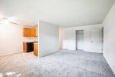 Located at 1998 NY-112 S in Coram, New York, this single-floor unit offers a straightforward, functional layout designed for comfortable everyday living. The living area is generously sized, featuring neutral gray carpeting, white walls, and a flush-mount ceiling light fixture. Multiple windows allow natural light to fill the space, while a wall-mounted HVAC unit and baseboard heating provide year-round climate control. A front door with adjacent windows adds additional brightness and connects directly to the living area.

The kitchen is semi-partitioned from the living space, creating a defined cooking area while maintaining an open feel. It is equipped with warm honey-toned wood cabinetry, a gas range with an over-the-range microwave, a black dishwasher, a full-size refrigerator, and light-toned tile flooring. The layout allows for efficient use of space throughout.

The bedroom is a well-proportioned room with the same neutral carpeting, ample wall space, and a closet with a standard rod for hanging storage. The bathroom includes a vanity with a white countertop sink and a mirrored medicine cabinet above, offering practical storage. The overall ambiance of the unit is clean, neutral, and move-in ready.