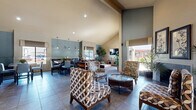 Located at 1522 E Southern Ave in Tempe, Arizona, this single-floor community space offers a thoughtfully designed open-plan layout that seamlessly connects a living area and dining room. The most striking architectural feature is the dramatic vaulted ceiling, which rises to impressive heights and enhances the overall sense of spaciousness throughout the interior.

The living area is comfortably furnished with patterned accent chairs, a plush sofa, and a substantial coffee table, all arranged to encourage conversation and relaxation. A wall-mounted flat-screen television anchors one end of the room, complemented by a wooden media console. Large windows dressed with tailored Roman shades invite generous natural light and provide views of the surrounding outdoor landscape.

The dining area features multiple high-top tables paired with upholstered counter-height chairs, illuminated by sleek pendant lights that hang above. Blue-toned accent wall panels add a layer of visual interest and depth, tying together the muted, neutral color palette found throughout the space. French doors along one wall provide direct outdoor access, further brightening the interior.

Throughout, large-format tile flooring and an abundance of tall indoor plants lend a clean, refreshing ambiance. The overall aesthetic is modern yet approachable, making this a welcoming and functional gathering space.