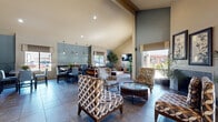 Located at 1522 E Southern Ave in Tempe, Arizona, this single-floor community space offers a thoughtfully designed open-plan layout that seamlessly connects a living area and dining room. The most striking architectural feature is the dramatic vaulted ceiling, which rises to impressive heights and enhances the overall sense of spaciousness throughout the interior.

The living area is comfortably furnished with patterned accent chairs, a plush sofa, and a substantial coffee table, all arranged to encourage conversation and relaxation. A wall-mounted flat-screen television anchors one end of the room, complemented by a wooden media console. Large windows dressed with tailored Roman shades invite generous natural light and provide views of the surrounding outdoor landscape.

The dining area features multiple high-top tables paired with upholstered counter-height chairs, illuminated by sleek pendant lights that hang above. Blue-toned accent wall panels add a layer of visual interest and depth, tying together the muted, neutral color palette found throughout the space. French doors along one wall provide direct outdoor access, further brightening the interior.

Throughout, large-format tile flooring and an abundance of tall indoor plants lend a clean, refreshing ambiance. The overall aesthetic is modern yet approachable, making this a welcoming and functional gathering space.
