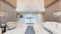 Located at 2971 Shell Road in Brooklyn, NY, this sophisticated lobby serves as a grand first impression for the building. The single-floor space is generously sized, offering a welcoming and polished environment defined by high-end finishes and thoughtful design.

The entryway features dramatic floor-to-ceiling glass panels flanking the main doors, flooding the space with natural light and framing views of the outdoors. Lush tropical plants are positioned at the entrance, adding a vibrant, organic touch to the modern aesthetic.

The flooring throughout is composed of large-format polished marble tiles in a classic white-and-gold veining pattern, lending a luxurious feel that extends across the entire hallway. A central reception desk, clad in matching marble with gold accents and branded signage, serves as the focal point of the lobby area. Flanking the reception are white upholstered sofas, offering comfortable seating for visitors.

Distinctive vertical wood slat accent walls line multiple sections of the space, introducing warmth and texture against the otherwise crisp, neutral palette. Built-in display niches within the wood paneling showcase decorative objects, adding character and detail. Recessed ceiling lighting and soaring ceiling heights further enhance the open, airy ambiance. A wall-mounted flat-screen display and a connecting hallway complete this refined, contemporary lobby.