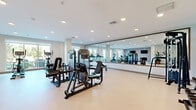 Located in Kissimmee, Florida, this fitness center offers a generously sized, single-floor exercise room designed to support a full range of workout routines. The space features a clean, modern aesthetic with neutral-toned walls, smooth light-colored flooring, and recessed lighting that creates a bright, energizing atmosphere throughout.

A floor-to-ceiling mirror spans an entire wall, visually expanding the room and providing exercisers with proper form feedback. Large windows along the perimeter allow natural light to filter in, enhancing the open and airy feel of the space.

The exercise room is comprehensively outfitted with a diverse array of equipment. Cardio offerings include treadmills, elliptical machines, and stationary bikes, while strength training is supported by multi-functional cable machines, weight benches, a dumbbell rack, a power rack, and various selectorized resistance machines. Wall-mounted flat-screen TVs provide entertainment during workouts.

Decorative touches, including framed fitness-themed artwork and potted plants, add warmth and personality to the space. A dedicated entry door ensures easy access, and the open floor plan provides ample room to move freely between equipment. The overall design balances functionality with a clean, professional ambiance suited for focused, effective training sessions.