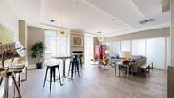 Located at 2401 Chicago Ave in Minneapolis, MN, this contemporary community lounge offers a well-appointed, single-floor layout designed for both relaxed socializing and casual gatherings. The open main area is anchored by light hardwood flooring and tray ceilings that lend a refined sense of height and structure throughout the space.

The lounge area is elegantly furnished with a plush gray sectional sofa, distinctive olive-toned swan chairs, and sleek glass-and-metal accent tables. Striking sputnik chandeliers serve as statement lighting fixtures, casting a warm glow over the room. Large floor-to-ceiling windows dressed with sheer curtains flood the interior with natural light and offer views of the surrounding streetscape. A fireplace adds a welcoming focal point to one wall, complemented by curated artwork and lush indoor greenery.

A bank of frosted glass sliding panels defines a semi-private zone within the lounge, providing flexible separation between areas. High-top bar tables with industrial-style black stools create a casual dining or work setting near the windows.

The kitchenette features dark wood cabinetry, stainless steel appliances including a full-size refrigerator and wine cooler, and a clean white countertop with an undermount sink. Thoughtful decorative touches, including bold abstract artwork and a vintage-style vending machine, give the space a vibrant, curated personality throughout.