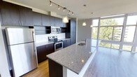 This single-floor space showcases a contemporary open-concept layout that seamlessly connects the kitchen and living areas. The kitchen is a standout feature, outfitted with dark espresso cabinetry that extends to the ceiling, providing generous storage capacity. A large granite-topped island with an integrated sink serves as both a functional workspace and a natural dividing point between the kitchen and living area. Stainless steel appliances, including a refrigerator, electric range, and built-in microwave, complement the modern aesthetic. Pendant lighting hangs above the island, while a track lighting system illuminates the kitchen workspace effectively. Light hardwood-style flooring runs throughout, adding warmth and continuity across the open layout. The living area is framed by expansive floor-to-ceiling windows that span an entire wall, flooding the interior with natural light and offering elevated views of surrounding trees and neighboring buildings. The overall ambiance is clean, bright, and modern, with a neutral color palette of whites and grays that enhances the sense of openness throughout the space.