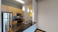 This single-floor space offers a thoughtfully designed open-concept layout that seamlessly connects the kitchen and living area. The kitchen is a standout feature, outfitted with warm honey-toned cabinetry, dark granite countertops, a mosaic tile backsplash, and a full suite of stainless steel appliances including a refrigerator, microwave, and electric range. A double-basin stainless steel sink is set into the peninsula counter, which also serves as a natural divider between the kitchen and the main living space. Stylish cylindrical pendant lights hang above, adding a modern touch. Light-toned hardwood-style flooring runs throughout the living area, complemented by exposed ductwork on the ceiling that lends an industrial character to the space. The bedroom is bright and airy, featuring the same light flooring, a sliding glass door fitted with vertical blinds that provides access to an outdoor area, and a side window that invites natural light. Ceiling fans provide comfort in both the living and bedroom areas. The bathroom is clean and functional, featuring large-format ceramic tile flooring, a full soaking tub with tiled surround, a coordinating granite-topped vanity with matching cabinetry, and a sleek sliding door entry.