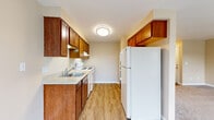 This single-story, one-bedroom, one-bathroom unit offers a thoughtfully arranged layout that maximizes its compact footprint. The open living room is finished with neutral-toned carpet and warm beige walls, creating a welcoming atmosphere. Large windows allow generous natural light to fill the space, with views of surrounding greenery. A sliding glass door in one configuration opens to an outdoor balcony, further enhancing the sense of openness.

The galley-style kitchen is efficiently designed, featuring rich warm-toned wood cabinetry on both walls, light countertops, a double sink, and white appliances including a refrigerator, electric range, and dishwasher. Light wood-style flooring runs throughout the kitchen, providing a clean contrast to the carpeted living areas.

The bedroom is comfortably sized, featuring carpet flooring, a window with outdoor views, and flush-mount lighting. Two closets provide practical storage throughout the unit. The bathroom includes a full tub-shower combination with white tile surround, wood-style flooring, and clean, functional fixtures. Overall, the unit presents a neutral, move-in-ready aesthetic with a consistent warm color palette throughout.