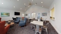 This well-appointed community lounge is designed to accommodate both relaxed gatherings and casual socializing. The space is anchored by dramatic vaulted ceilings that rise to a sharp peak, fitted with a ceiling fan and recessed lighting that floods the room with a bright, airy atmosphere. White walls are accented by a bold mustard-yellow feature wall surrounding a teal-tiled fireplace, which serves as the room's visual centerpiece. Flanking arched windows allow ample natural light to filter in, enhancing the open and welcoming feel throughout.

The layout is divided into distinct functional zones. A lounge area features plush gray sofas, blue wingback chairs, and terracotta accent chairs arranged around glass and wood coffee tables, encouraging comfortable conversation. Nearby, a dining zone is outfitted with industrial-style metal chairs and white-topped tables, ideal for group use. A wall-mounted flat-screen television adds an entertainment focal point to the seating area.

Bold, colorful artwork is displayed throughout, contributing a modern and vibrant character. Geometric-patterned carpeting in dark tones grounds the space and adds visual texture underfoot. An open pass-through connects to a kitchenette visible in the background, extending the space's utility for events and gatherings. Frosted glass doors provide access to additional rooms, rounding out this versatile community facility.