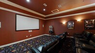 This private home theater is designed to deliver an immersive cinematic experience from the comfort of home. The room is finished in rich, deep terracotta-red walls that set a dramatic, theater-inspired atmosphere, complemented by white crown molding that frames the tray ceiling above. Recessed lighting and warm wall sconces provide layered ambient illumination, enhancing the room's intimate ambiance.

The centerpiece of the space is a large, wall-mounted projection screen positioned at the front of the room, flanked by in-wall speaker panels that support a full surround sound setup. Multiple rows of plush, black leather recliner seats with built-in cup holders are arranged to provide clear sightlines to the screen from every position, comfortably accommodating a generous number of viewers.

Underfoot, a vibrant patterned carpet with colorful motifs adds a playful, classic cinema feel to the space. Framed movie posters — including recognizable blockbuster titles — adorn the rear and side walls, reinforcing the theatrical character of the room. A traditional-style popcorn machine on a stand adds an authentic concession-stand touch, completing the full moviegoing experience. This thoughtfully designed space offers every detail needed for private, high-quality film screenings.