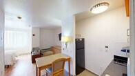 This compact studio in New Orleans, LA, offers a well-organized single-floor layout that efficiently incorporates a bedroom area, full kitchen, and bathroom. The living and sleeping space is defined by a bold orange accent wall that adds warmth and visual energy to the room, complementing the light hardwood-style flooring throughout. A full-size bed with a wooden headboard, wall-mounted lighting, and a bedside table furnish the sleeping area, while a seating area with an upholstered chair and ottoman provides a comfortable secondary space. A large window with vertical blinds allows natural light to brighten the room, and a wall-mounted flat-screen television adds convenience.

The kitchen is neatly arranged with white upper cabinetry accented by a vibrant orange soffit, dark countertops, a stainless steel sink, a microwave, and a full-size refrigerator, providing adequate functionality for everyday meal preparation. A small dining table with chairs is positioned nearby, creating a defined eating area.

The bathroom features light tile flooring, a tub and shower combination with a bright orange curtain, a toilet, a wall-mounted medicine cabinet, and an accessible grab bar. The overall ambiance is functional, colorful, and efficiently designed for comfortable single-occupancy living.
