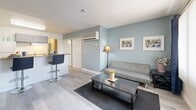 Located at 5025 Nellis Oasis Lane in Las Vegas, NV, this single-floor studio unit offers a well-organized and efficiently designed layout. The open-concept living area is finished with light wood-style flooring, soft blue-toned accent walls, and a textured ceiling, creating a calm and cohesive atmosphere throughout the space. A large window dressed with navy grommet curtains and sheer panels allows natural light to fill the room, enhancing its airy feel. A wall-mounted HVAC unit provides climate control directly within the living area. The kitchenette is positioned adjacent to the living space and features a breakfast bar with bar stool seating, a countertop workspace, and a compact sink — making it both functional and space-conscious. A sliding closet door provides built-in storage within the main living area. The bathroom is equipped with a full soaking tub and shower combination accented by a bold blue and white patterned curtain, a gray vanity with a warm-toned countertop, an integrated sink, and a large mirror. Tile flooring completes the bathroom's clean, utilitarian design. Overall, the unit presents a practical and neatly appointed living environment suited for single occupancy in the Las Vegas area.