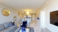 This single-floor residence showcases a cohesive contemporary design carried throughout its open-concept layout. The living area serves as the heart of the home, finished with large-format marble-look porcelain tile flooring that extends seamlessly across the entire unit. A plush gray sofa anchors the space, accented by navy blue throw pillows and a sleek white coffee table. A cluster of decorative circular mirrors in silver and gold tones adds visual interest to the walls. Floor-to-ceiling sliding glass doors flood the living room with natural light and open onto a private balcony, offering elevated views of the surrounding cityscape.

The dining area transitions naturally from the living space and features a glass-top table with modern white chairs beneath a sculptural pendant light fixture. The adjacent kitchen is well-appointed with dark wood cabinetry, light quartz countertops, stainless steel appliances including a full-size refrigerator, and track lighting overhead.

The bedroom is a calm, neutral retreat with a low-profile platform bed, a textured accent wall, and a window framing outdoor views. A wall-mounted flat-screen television and a mirrored closet complete the room. The bathroom features marble-look wall tile in the shower-tub combination, a dark wood vanity with a white countertop, and polished chrome fixtures throughout.