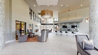 This contemporary clubhouse makes a striking first impression with its soaring double-height ceilings, floor-to-ceiling glass walls, and an abundance of natural light that floods the interior. Stately stone-clad columns anchor the open-plan layout, while large-format light tile flooring runs seamlessly throughout, enhancing the sense of spaciousness.

The welcoming reception area features a sleek, modern desk accented by warm wood tones and curated artwork, setting an elegant tone from the moment of arrival. Adjacent lounge areas are thoughtfully arranged with high-back upholstered chairs, bold area rugs in rich terracotta hues, and sculptural pendant lighting — including a statement drum pendant with warm wood detailing — creating intimate yet open gathering spaces.

A fully appointed community kitchen stands out with dramatic blue mosaic tile backsplash, stainless steel appliances, dark granite countertops, a large central island with bar seating, and pendant lighting — all framed by expansive windows with direct views to an outdoor pool area.

Additional seating vignettes offer versatile lounge configurations, featuring vibrant striped upholstery and artistic ceiling installations. A glass-enclosed conference or meeting room provides a private workspace option within the larger communal setting. Throughout, curated art pieces and lush greenery contribute to a polished, resort-inspired ambiance.