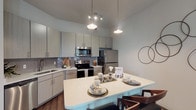 This single-floor residence offers a thoughtfully designed open-concept layout with contemporary finishes throughout. The kitchen serves as a central hub, featuring light gray flat-front cabinetry, a tiled backsplash, stainless steel appliances including a refrigerator, range, microwave, and dishwasher, and a large island with a crisp white countertop that provides ample seating and workspace. Pendant lighting and recessed track lights illuminate the space with a modern, layered glow. Rich dark hardwood-style flooring flows through the kitchen and living areas, adding warmth and contrast to the neutral color palette. The living room is generously proportioned, offering large windows that invite natural light, a ceiling fan, and direct sightlines to the bedroom, creating an airy, connected feel. The bedrooms feature plush carpeting and large windows with street views. One bedroom includes a dramatic tufted wall panel headboard feature and direct access to an en-suite bathroom. The bathrooms are finished with large-format stone-look tile on the walls and floors, soaking tubs, and vanities topped with granite countertops. Overall, the space presents a cohesive modern aesthetic with quality materials and functional design throughout.