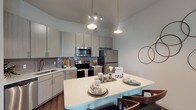 This single-floor residence offers a thoughtfully designed open-concept layout with contemporary finishes throughout. The kitchen serves as a central hub, featuring light gray flat-front cabinetry, a tiled backsplash, stainless steel appliances including a refrigerator, range, microwave, and dishwasher, and a large island with a crisp white countertop that provides ample seating and workspace. Pendant lighting and recessed track lights illuminate the space with a modern, layered glow. Rich dark hardwood-style flooring flows through the kitchen and living areas, adding warmth and contrast to the neutral color palette. The living room is generously proportioned, offering large windows that invite natural light, a ceiling fan, and direct sightlines to the bedroom, creating an airy, connected feel. The bedrooms feature plush carpeting and large windows with street views. One bedroom includes a dramatic tufted wall panel headboard feature and direct access to an en-suite bathroom. The bathrooms are finished with large-format stone-look tile on the walls and floors, soaking tubs, and vanities topped with granite countertops. Overall, the space presents a cohesive modern aesthetic with quality materials and functional design throughout.