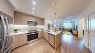 This thoughtfully designed single-floor residence combines modern aesthetics with functional living. The open-concept kitchen is a standout feature, outfitted with light wood cabinetry extending to the ceiling, a subway tile backsplash, white quartz countertops, and a full suite of stainless steel appliances including a range, built-in microwave, and dishwasher. A peninsula with a undermount sink provides additional prep space and naturally connects the kitchen to the rest of the living area. Track lighting and elegant pendant fixtures illuminate the space throughout. The dining area offers an intimate setting with clean white walls, ideal for displaying artwork. The living room is bright and welcoming, benefiting from large windows that invite natural light and feature a hardwood floor that runs continuously through the main living spaces. The bedroom is a serene retreat, offering access to both a private bathroom and a dedicated walk-in closet with built-in shelving. The bathroom features large-format marble-look tile on both the floors and tub surround, creating a clean, spa-like atmosphere. Overall, the residence presents a cohesive modern design with warm wood tones, neutral walls, and refined finishes throughout.